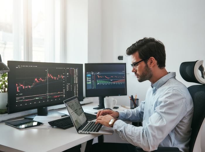 A man types into a laptop, there are two screens on a desk showing financial information